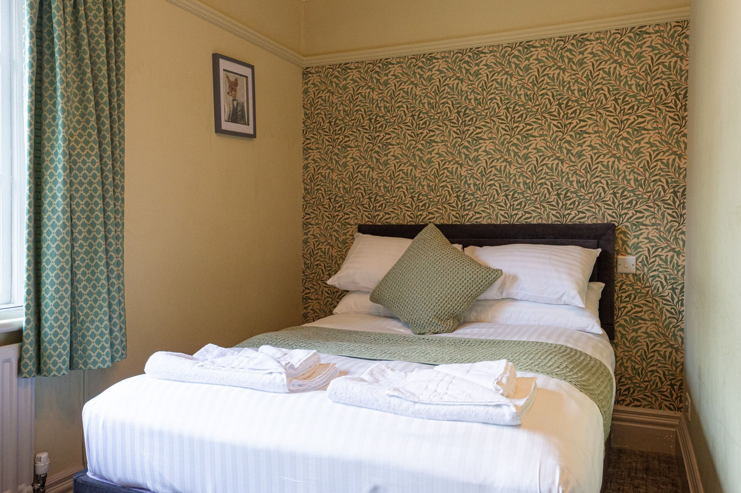 A neatly made bed with white linens and a green throw blanket, a pillow, and a folded towel on the bed.