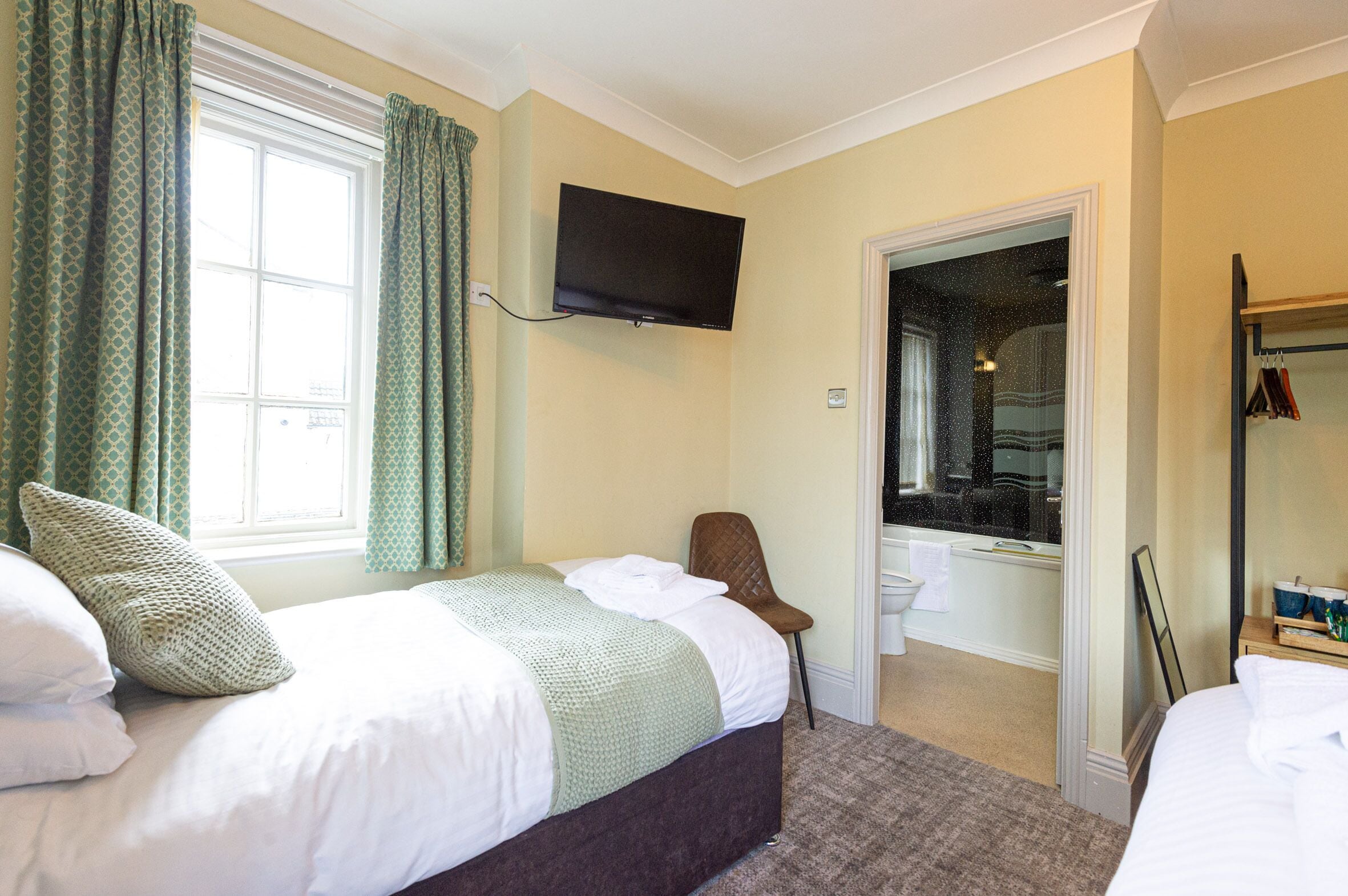 A hotel room with a bed, a chair, a television, a bathroom, and a coat rack.