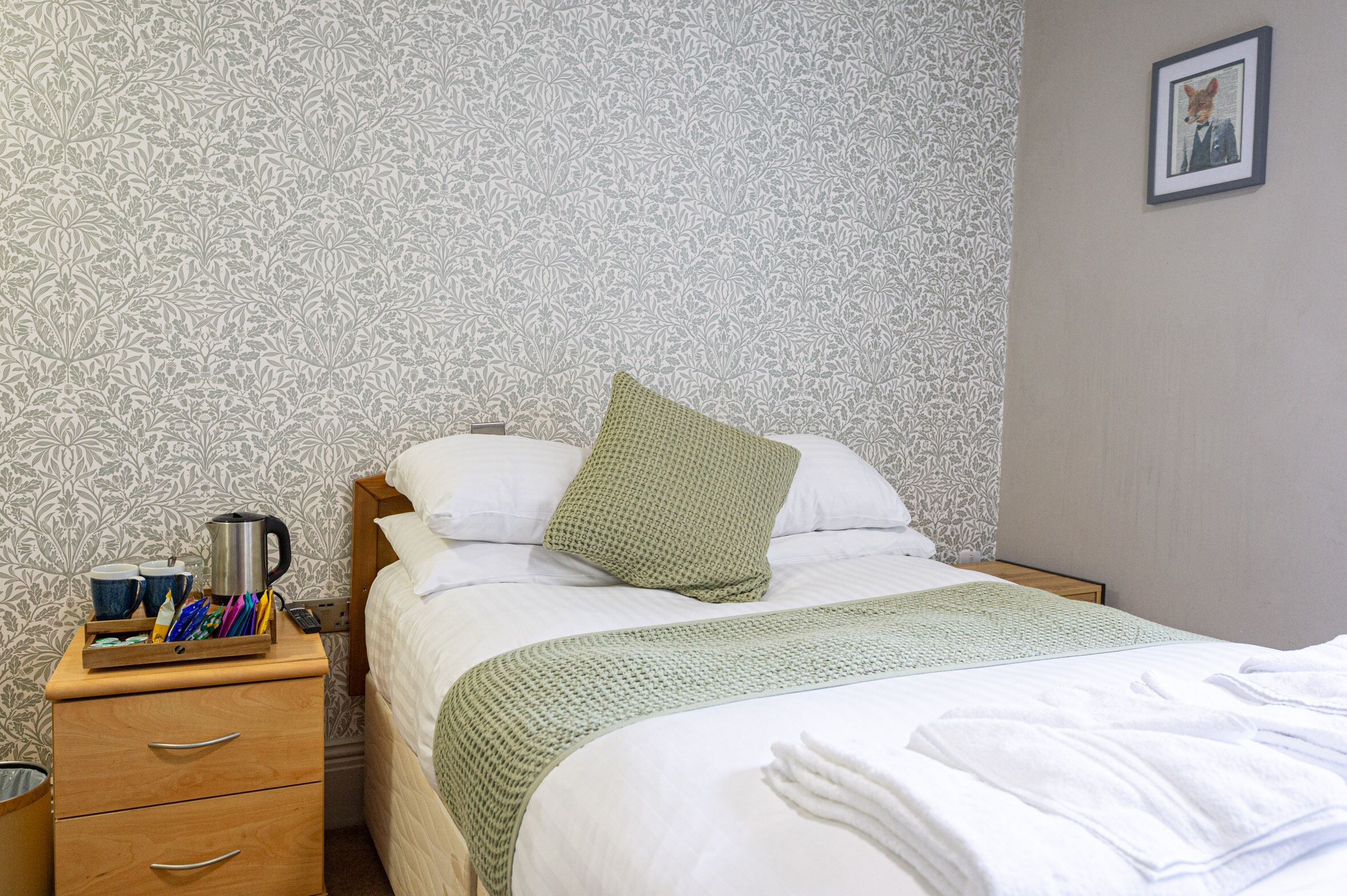 A neatly made bed with a green pillow, a bedside table with a kettle, cups, and a tray of pens, and a patterned wallpaper.