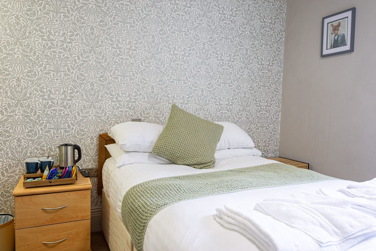 A neatly made bed with a green pillow, a bedside table with a kettle, cups, and a tray of pens, and a patterned wallpaper.