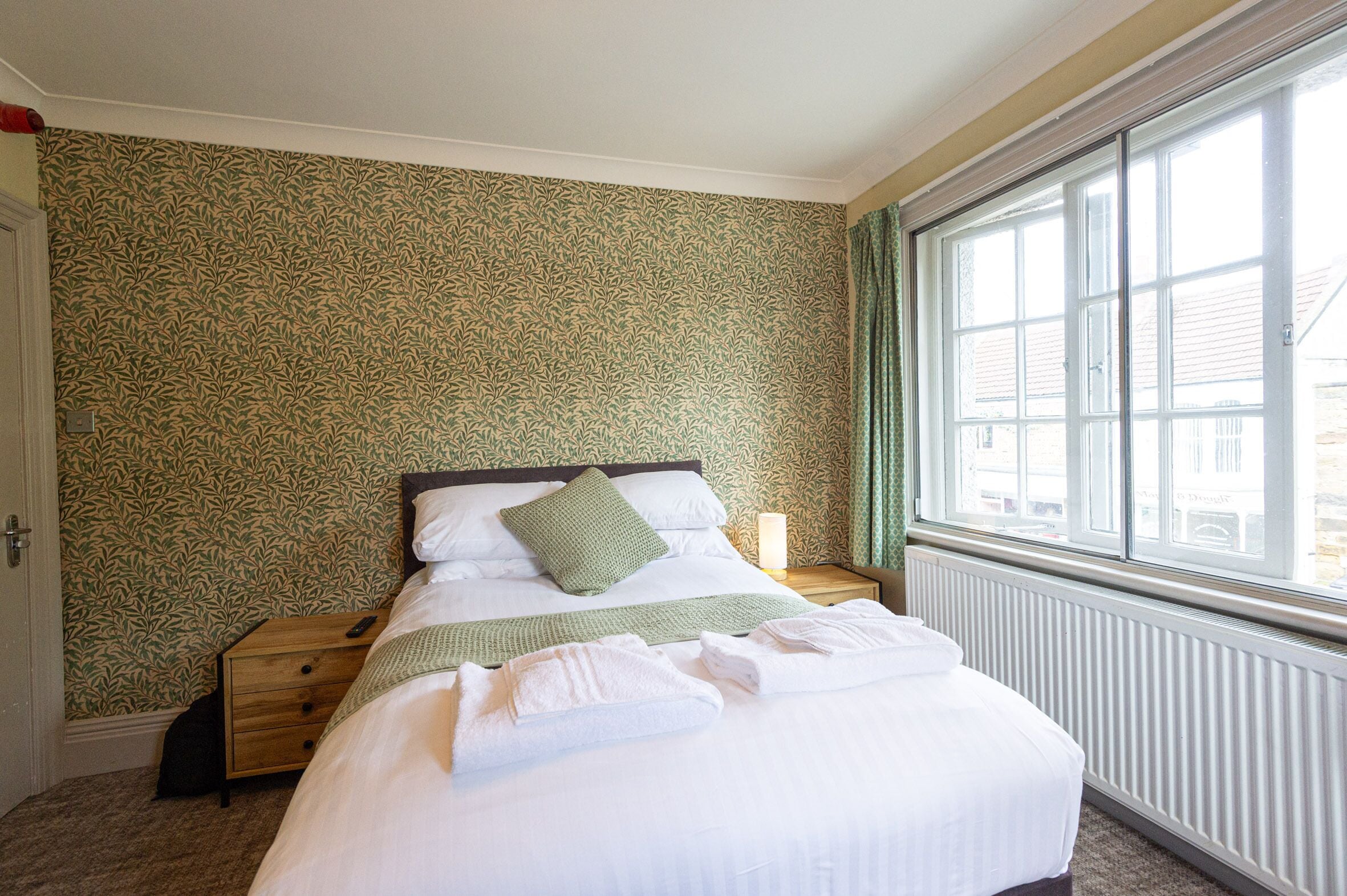 A neatly made bed with white linens and a green throw, a bedside table with a lamp, and a window with a view of buildings outside.