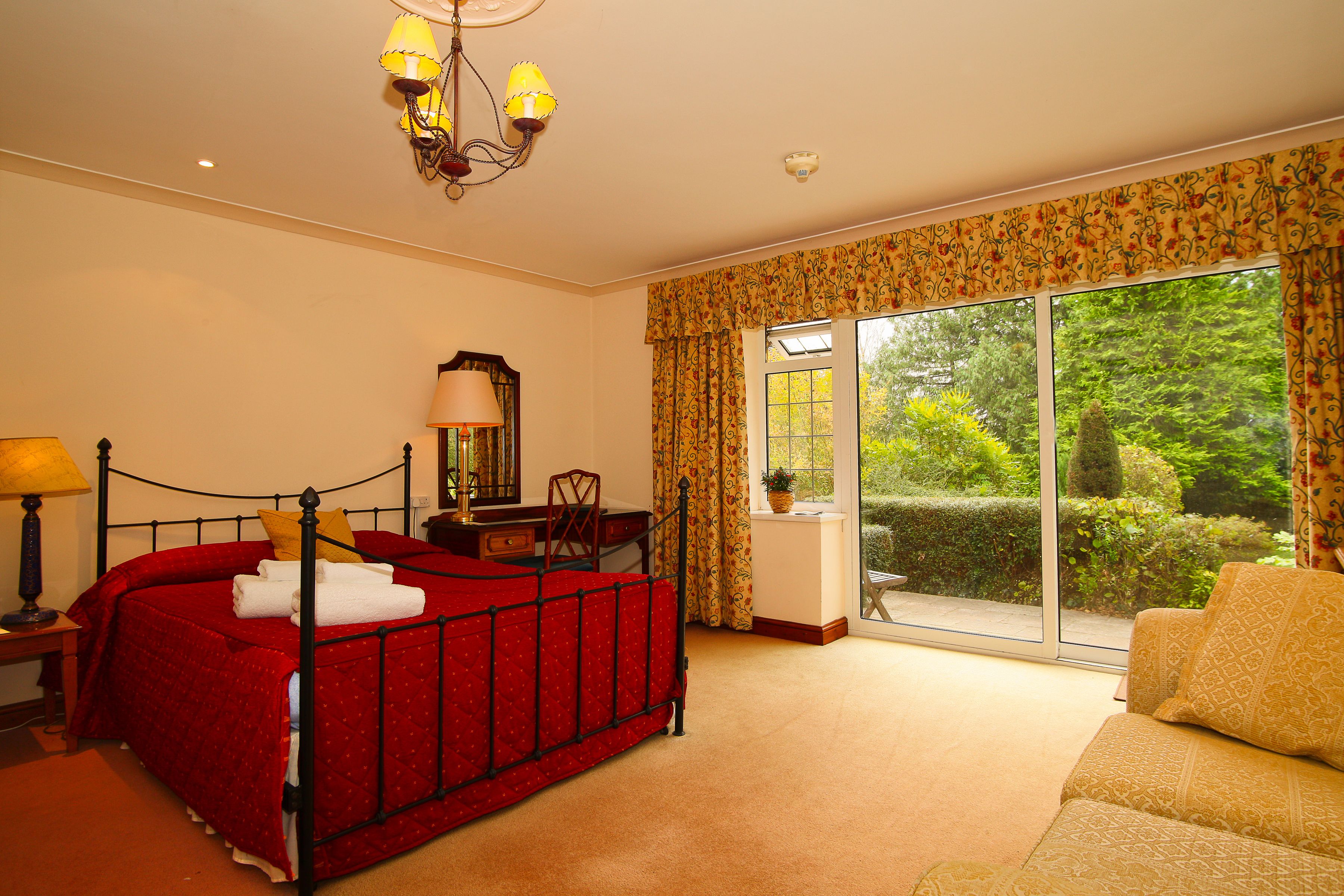 A bedroom with a large bed, a chair, a desk, and a sofa. There is a window with curtains and a view of greenery outside.