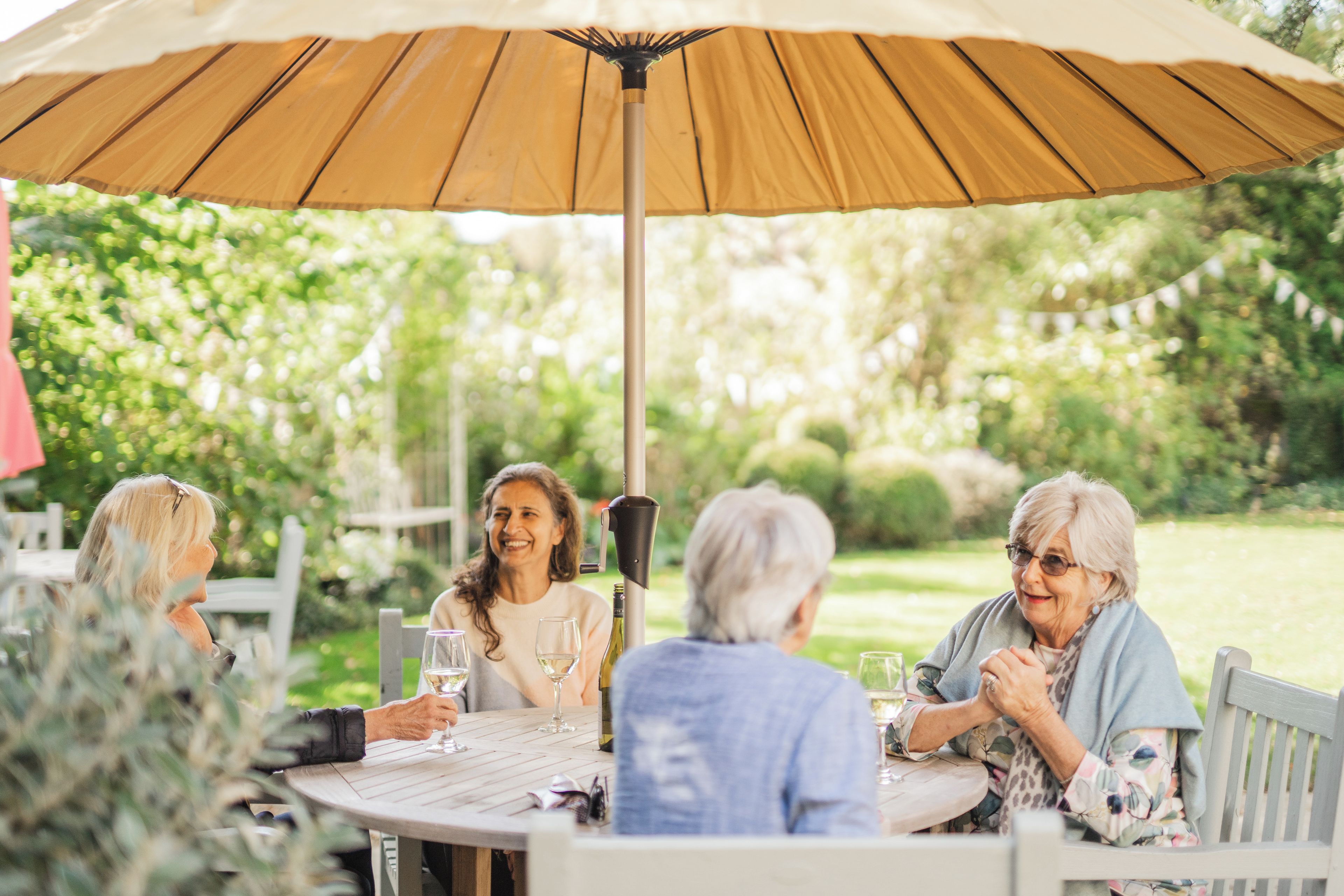 A group of people seated around a round table with a large umbrella overhead, in an outdoor setting with greenery in the background.