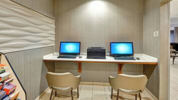 A modern office space with two desks, each with a laptop and a printer. There are two chairs at each desk, and a bookshelf filled with books is visible to the left.