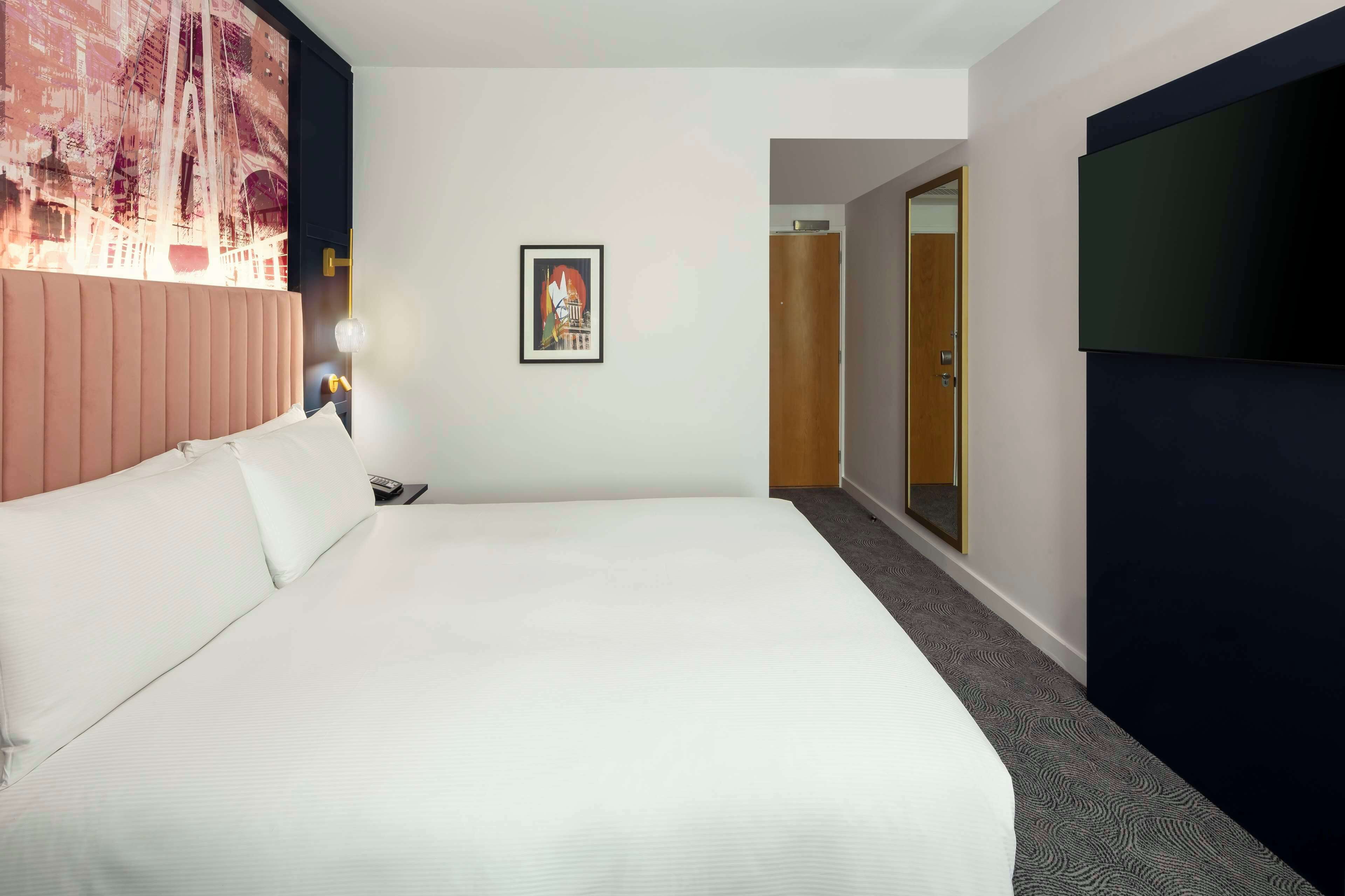 Habitación de hotel con una cama grande, un cuadro en la pared, un televisor a la derecha y una puerta que da a otra habitación.