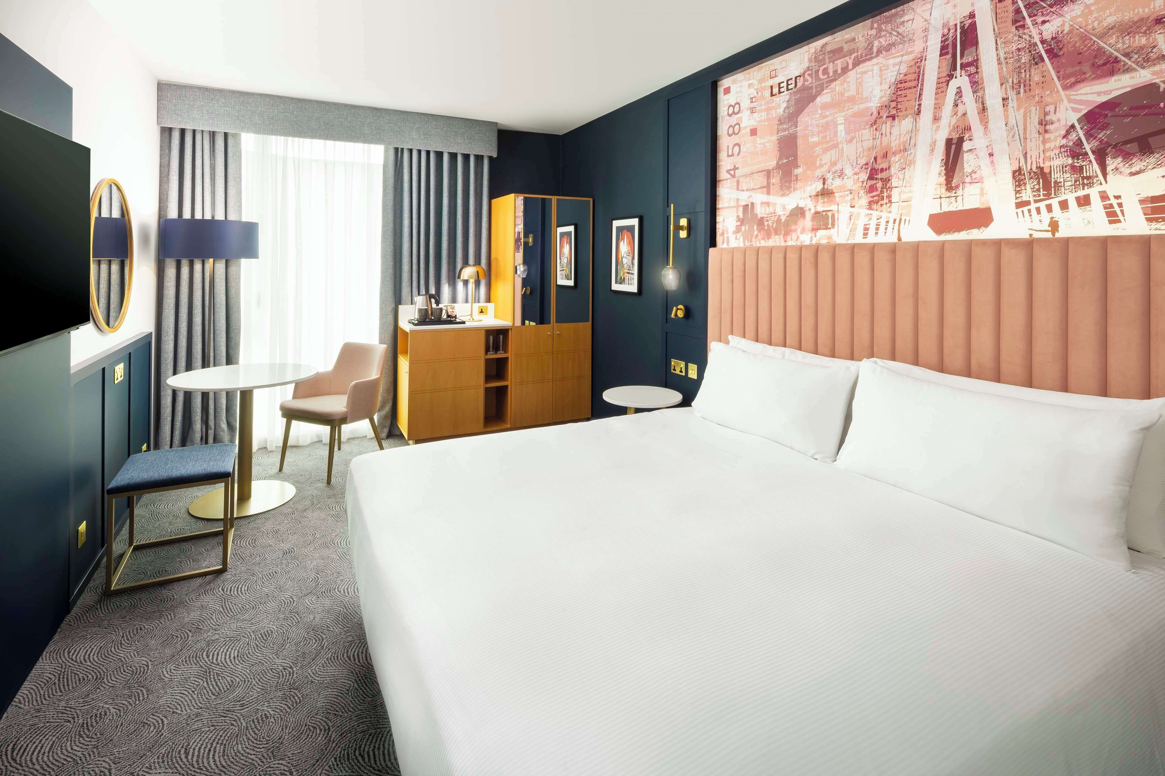 Habitación de hotel con una cama grande, un escritorio, una silla, un televisor y un mural con vista a la ciudad.