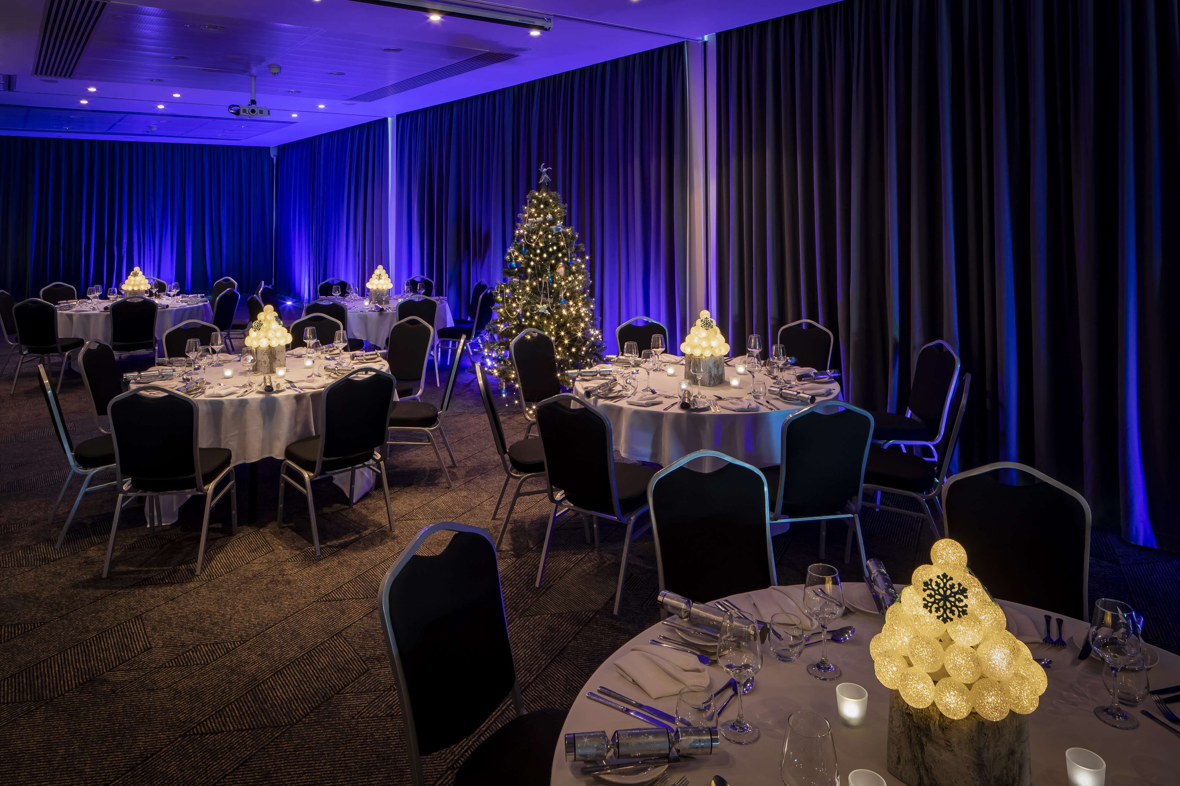A banquet hall with round tables set for dining, a decorated Christmas tree, and blue ambient lighting.