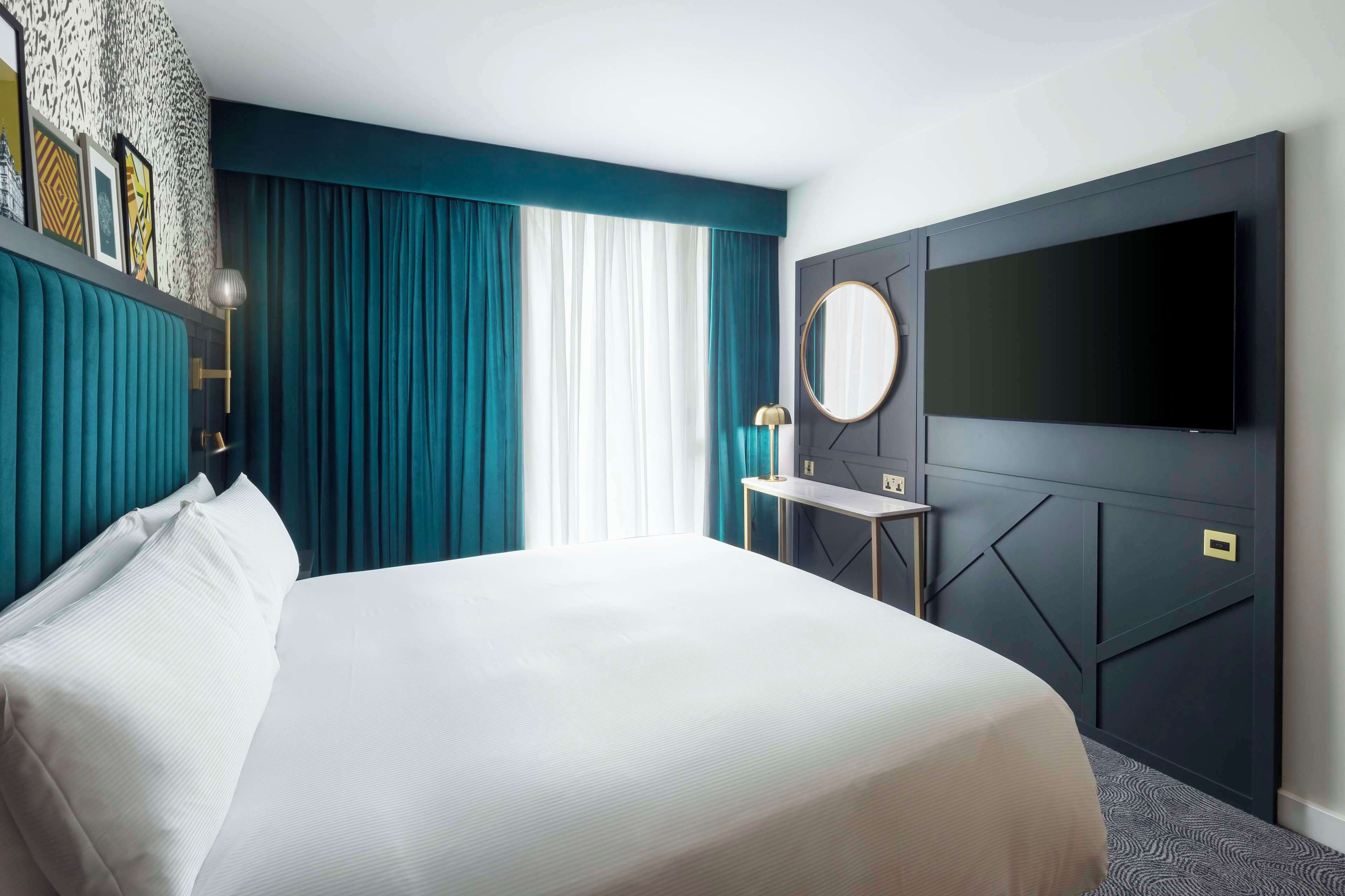 A hotel room with a large bed, a flat-screen TV mounted on the wall, a small table with a lamp, and a window with teal curtains.