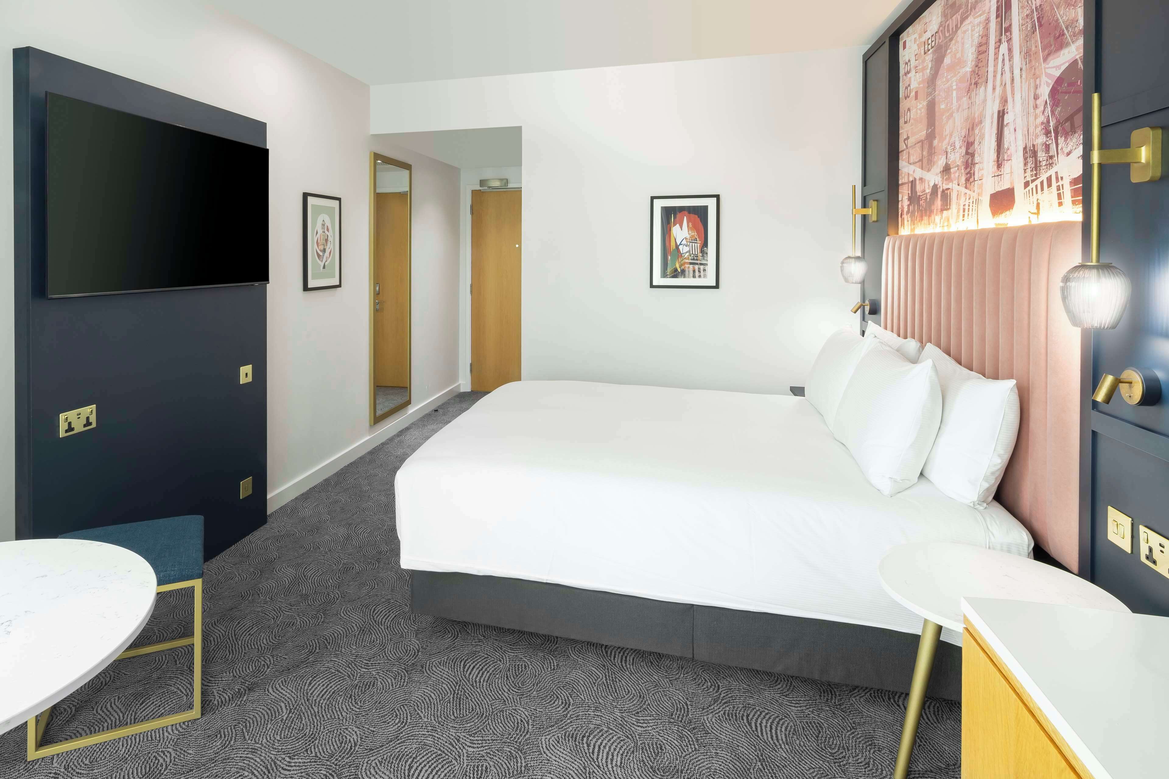 Quarto de hotel com uma cama grande, televisão de tela plana na parede, uma mesa redonda pequena e uma porta que dá para outro cômodo.
