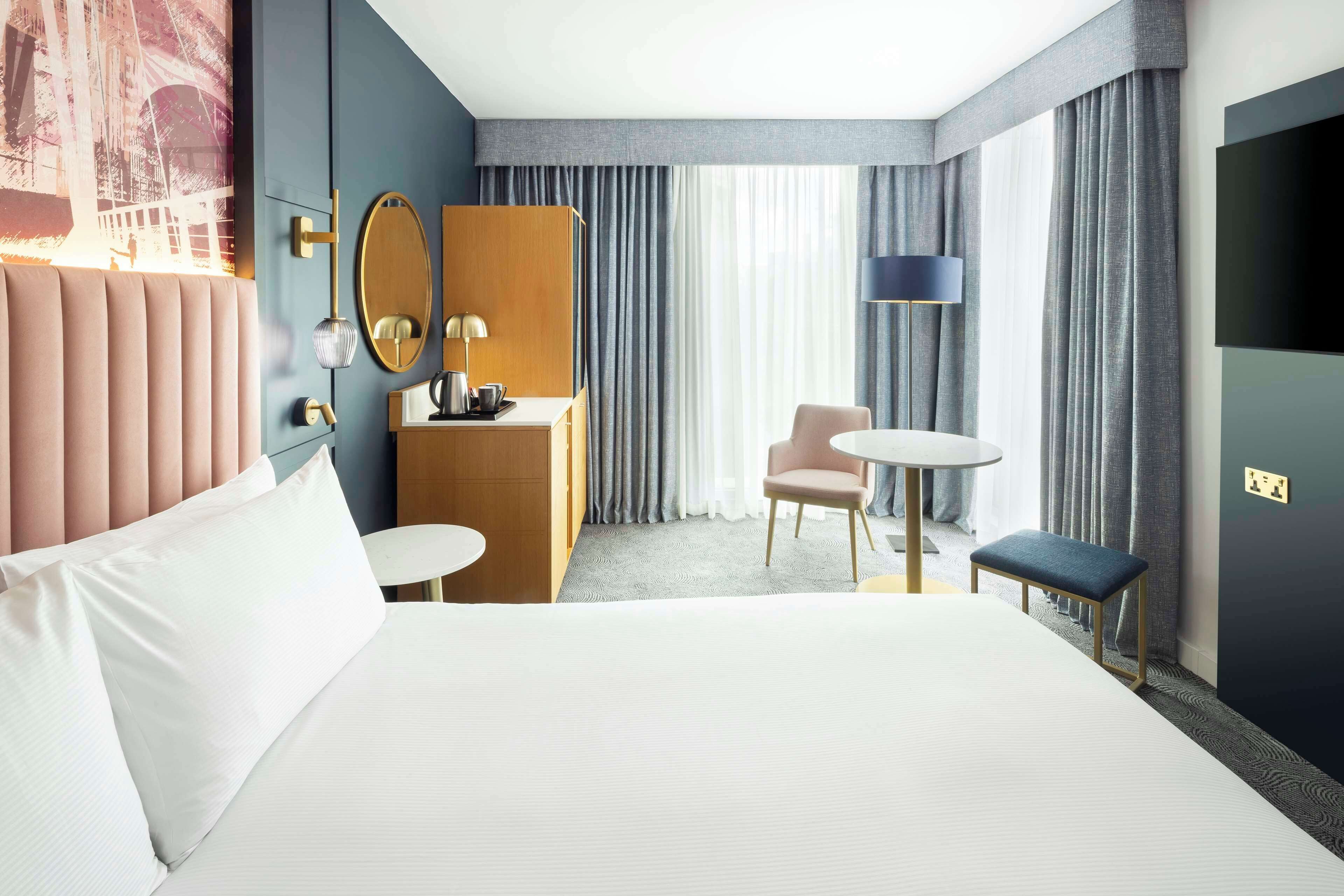 A modern hotel room with a large bed, a desk, a chair, and a small table.