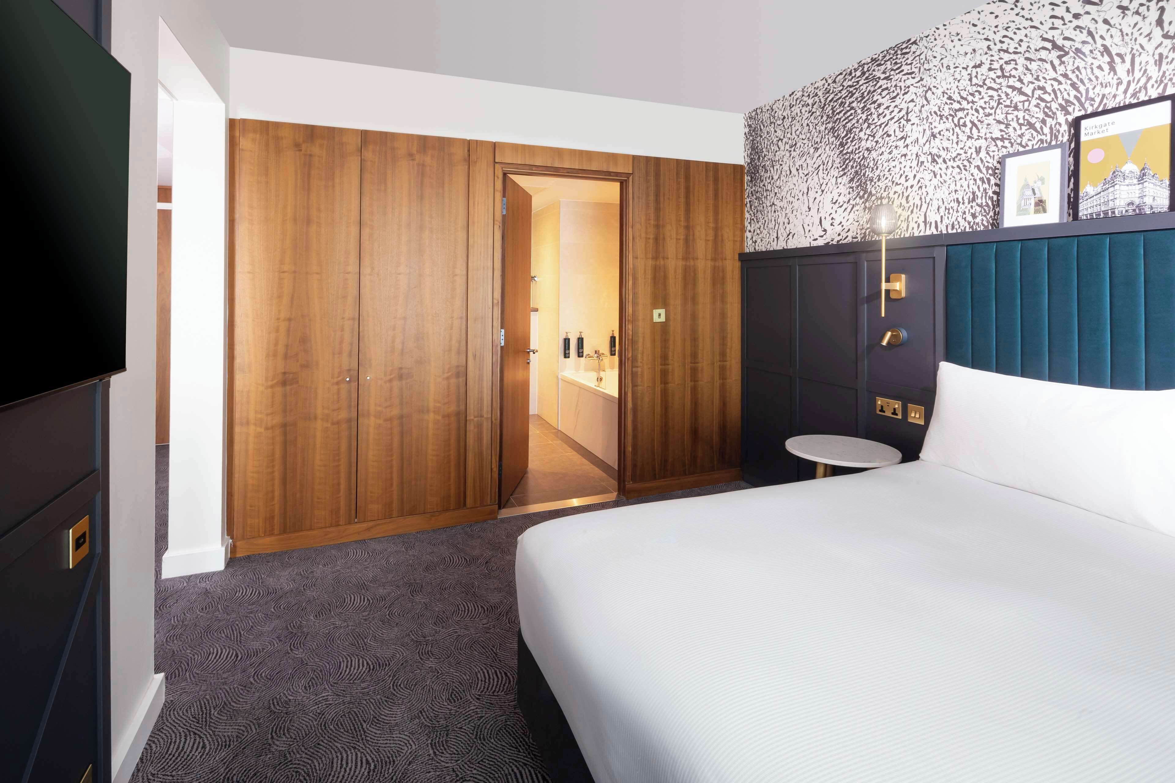 A hotel room with a bed, a TV, a wardrobe, a small round table, and a bathroom visible through an open door.