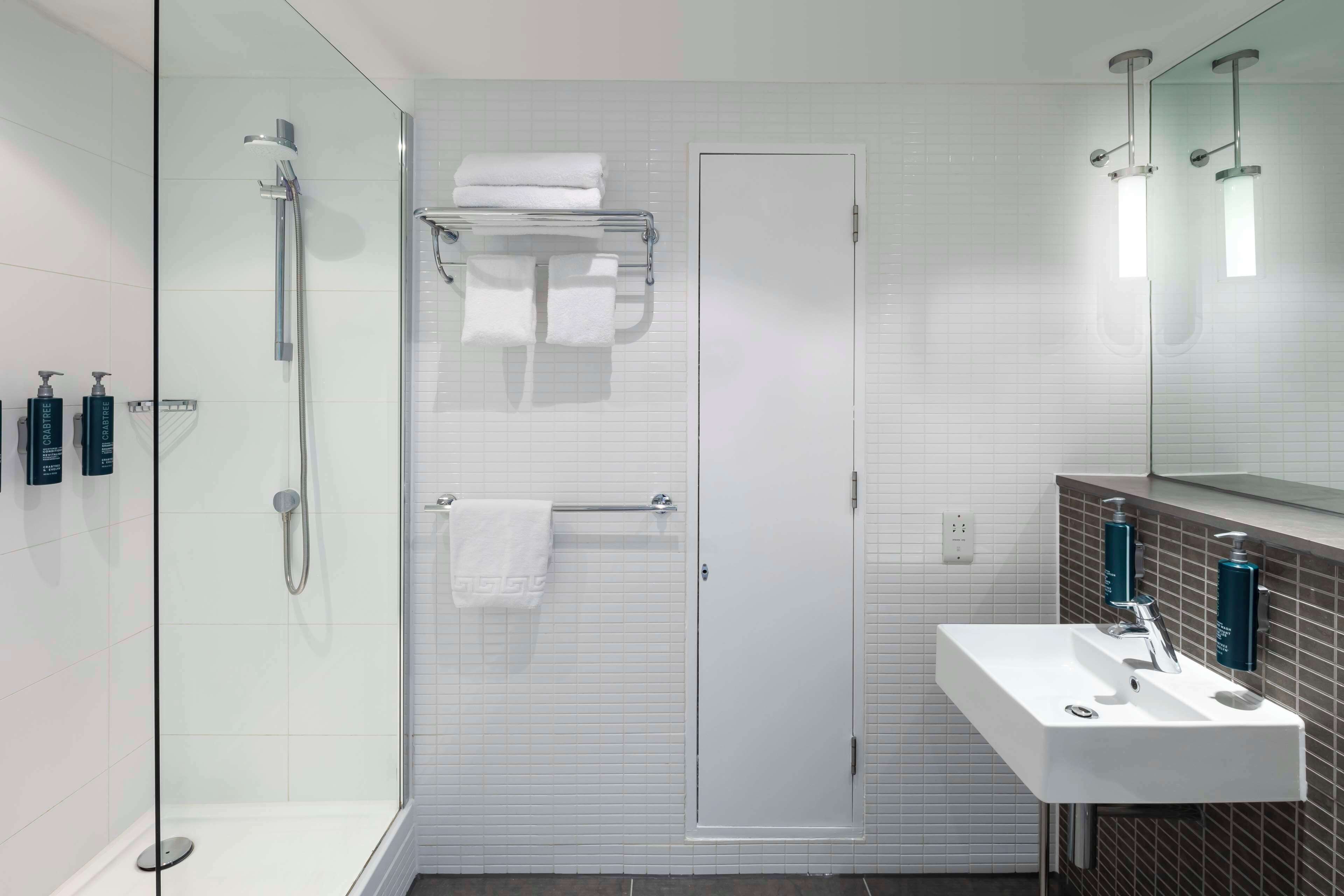 Une salle de bain moderne avec une douche à l’italienne en verre, un lavabo blanc avec un miroir au-dessus et des articles de toilette fixés au mur.