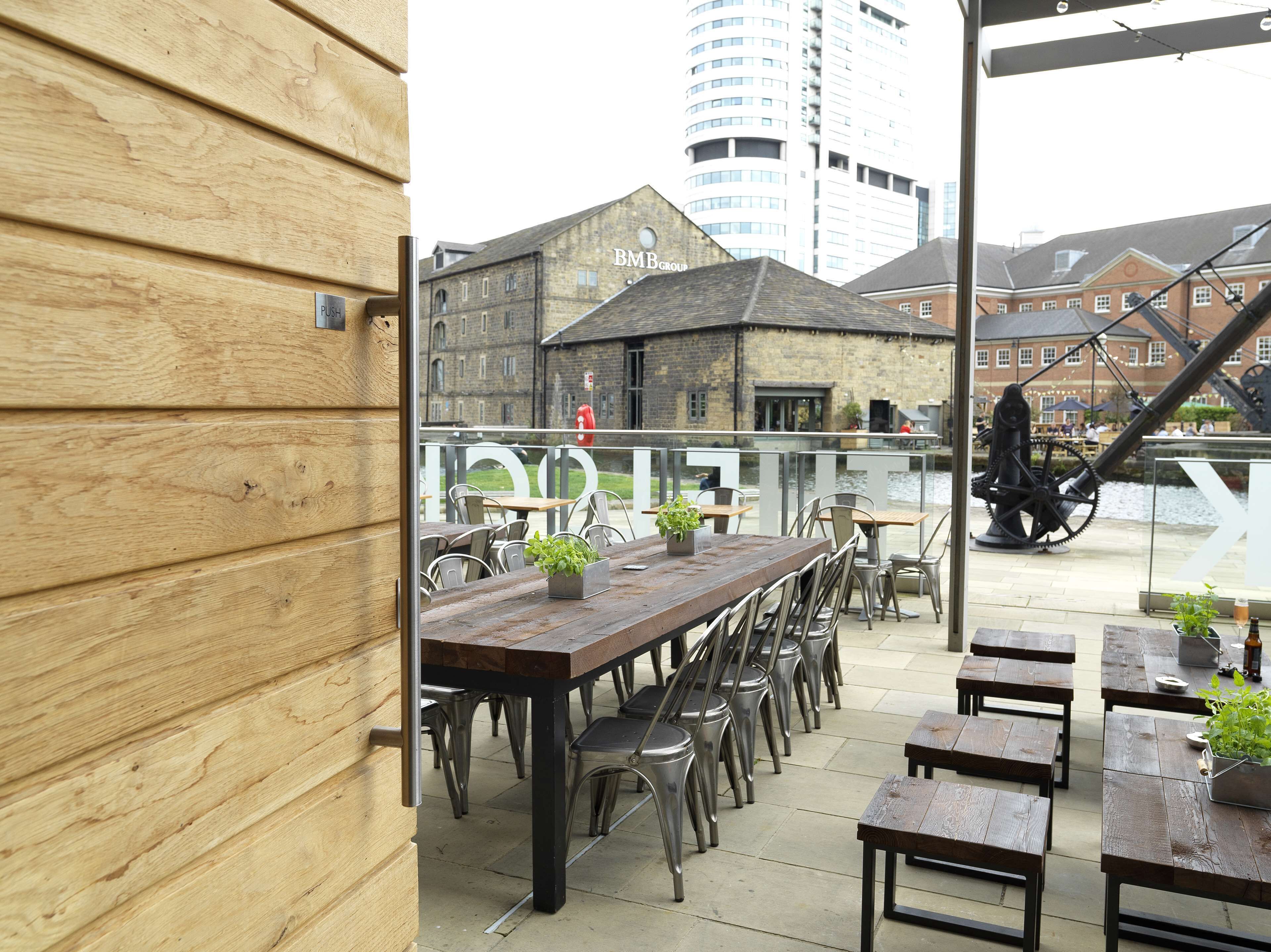 An outdoor seating area with wooden tables and metal chairs, overlooking a river with industrial buildings in the background.
