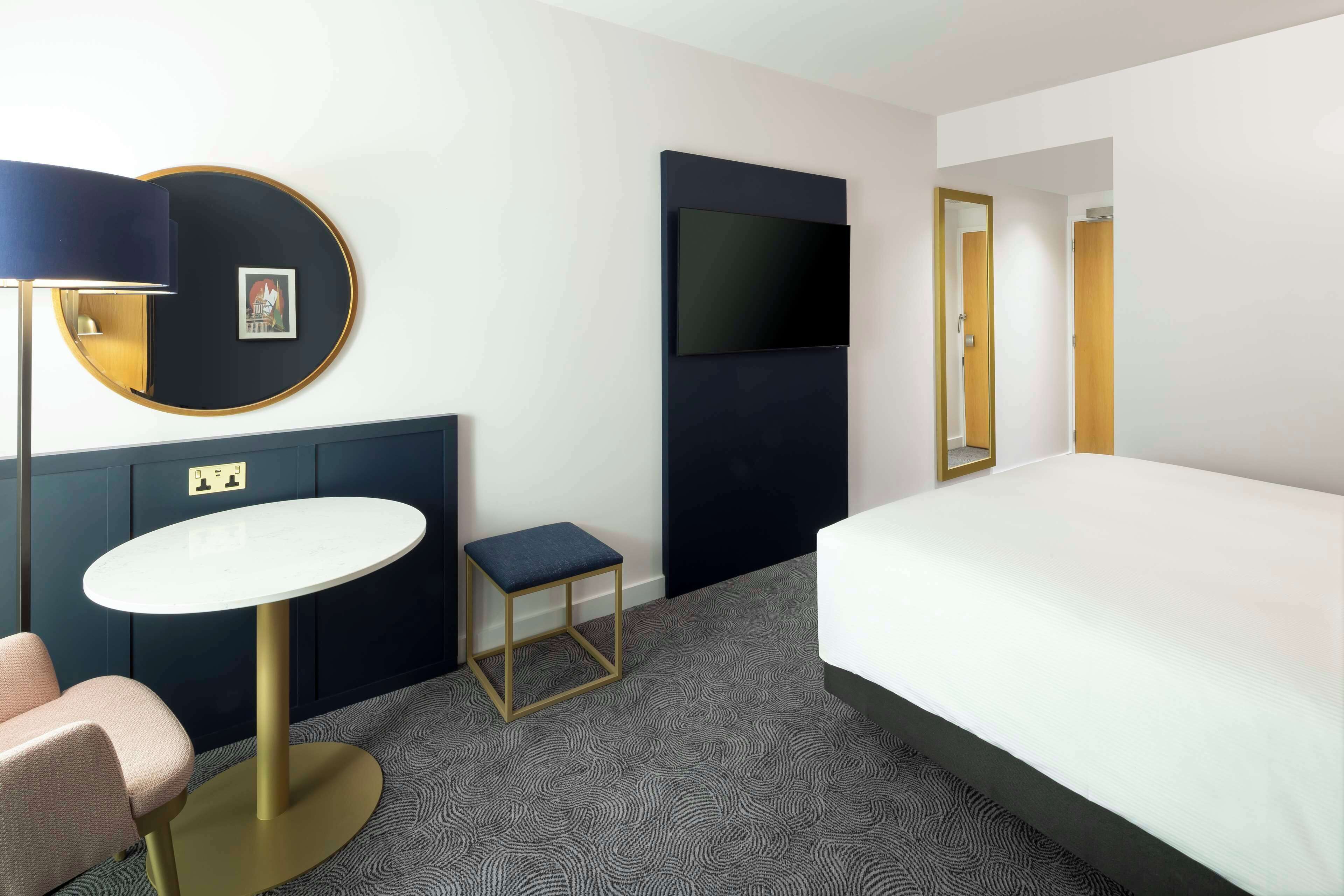 A hotel room with a bed, a round table, a small stool, a wall-mounted TV, and a door.