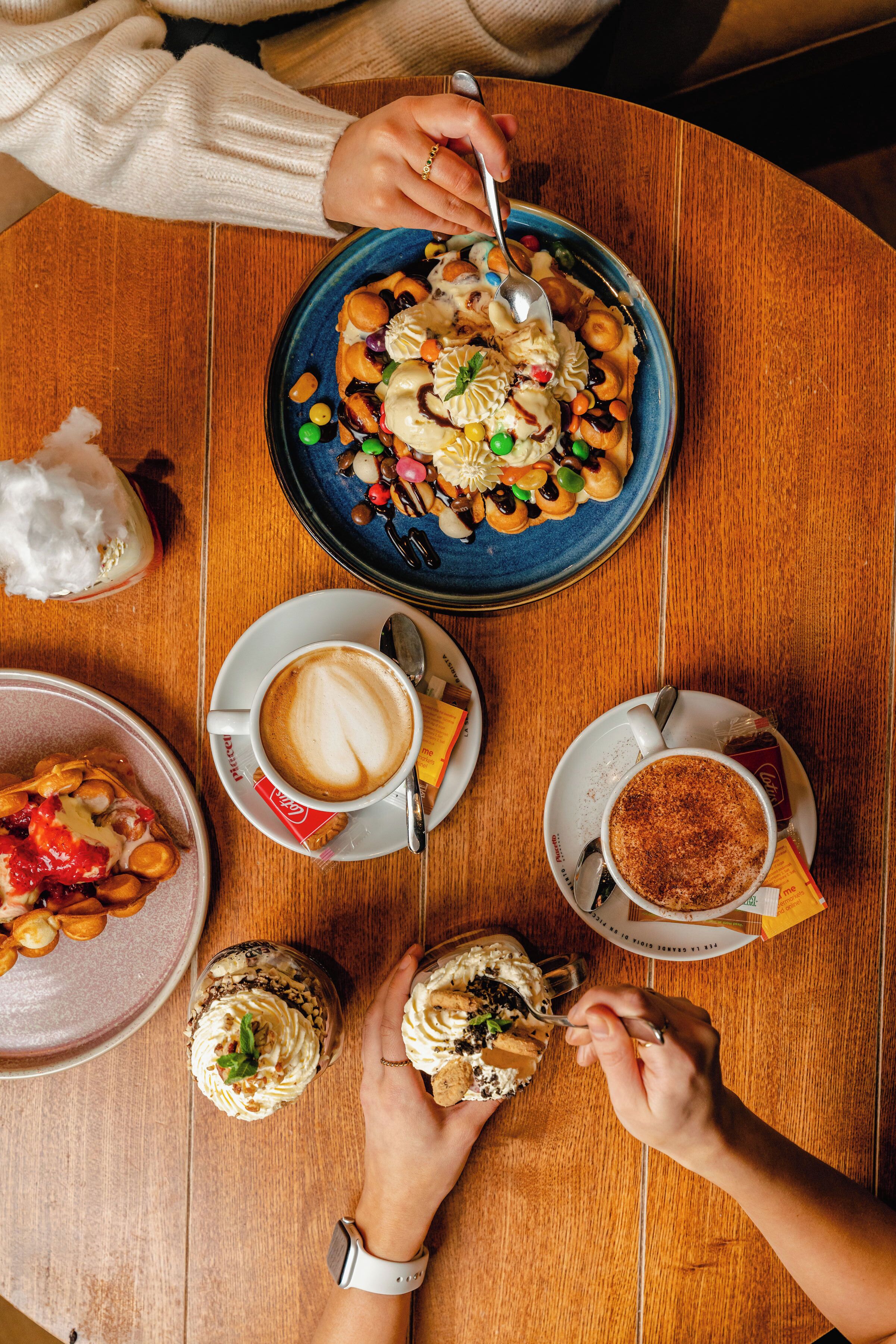 Una mesa de madera con diversos alimentos y bebidas, como pasteles, una taza de café y un plato de frutos secos y semillas.