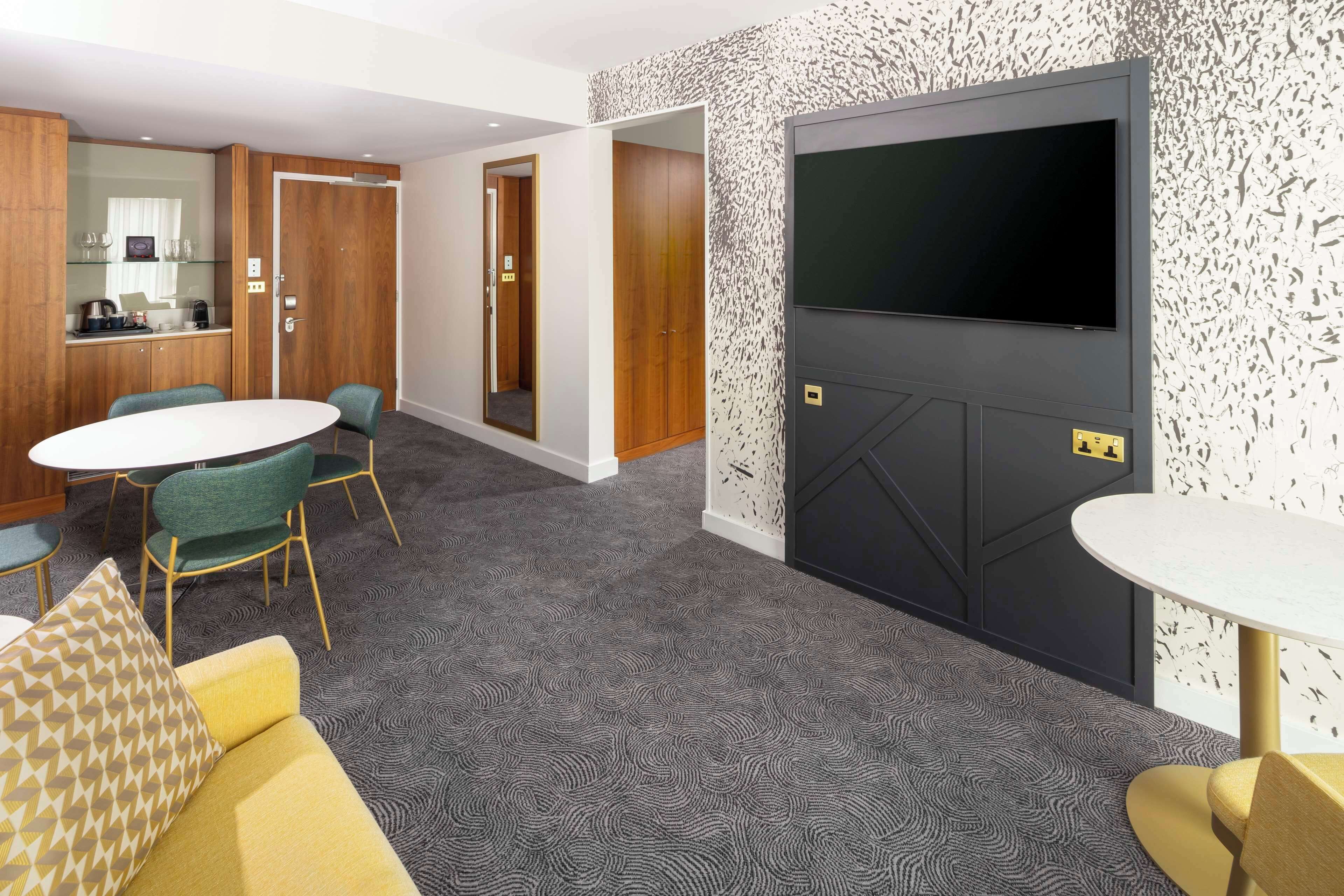 A hotel room with a large flat-screen TV, a dining area with a round table and chairs, a sofa with patterned cushions, and a carpeted floor.