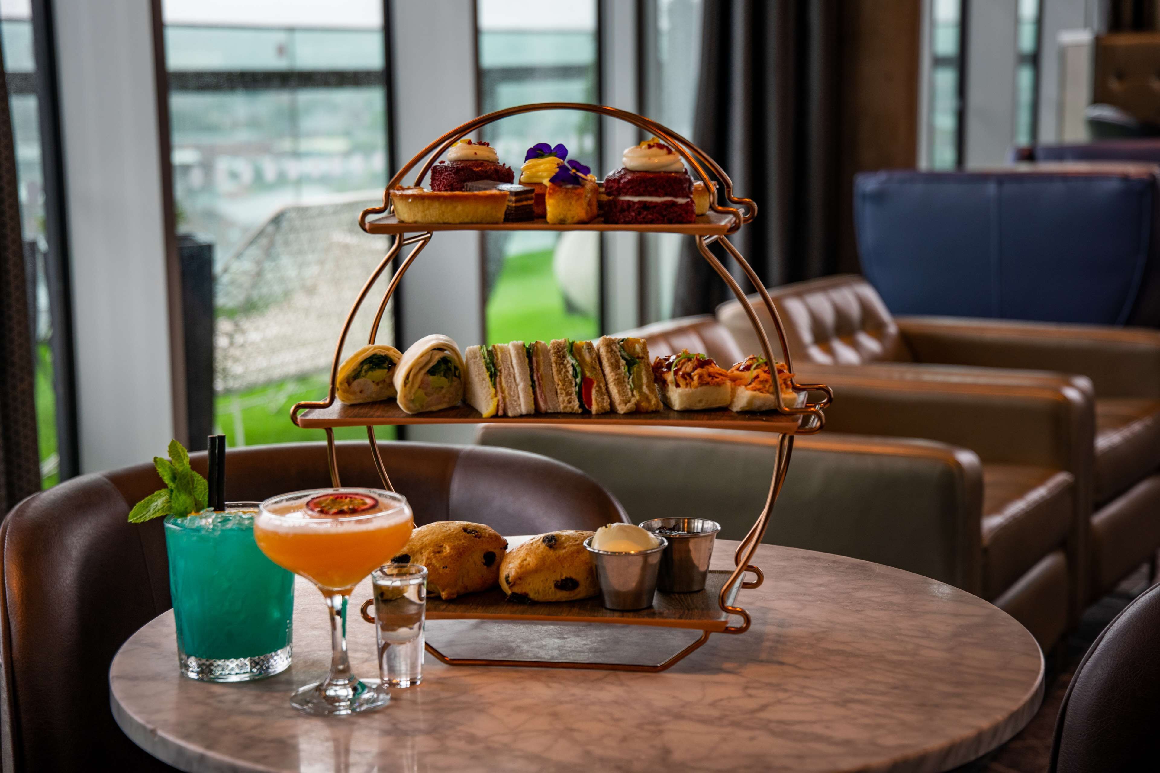 A tiered dessert tray with various pastries and sandwiches on a table in a seating area with leather chairs and a view of a green landscape through large windows.