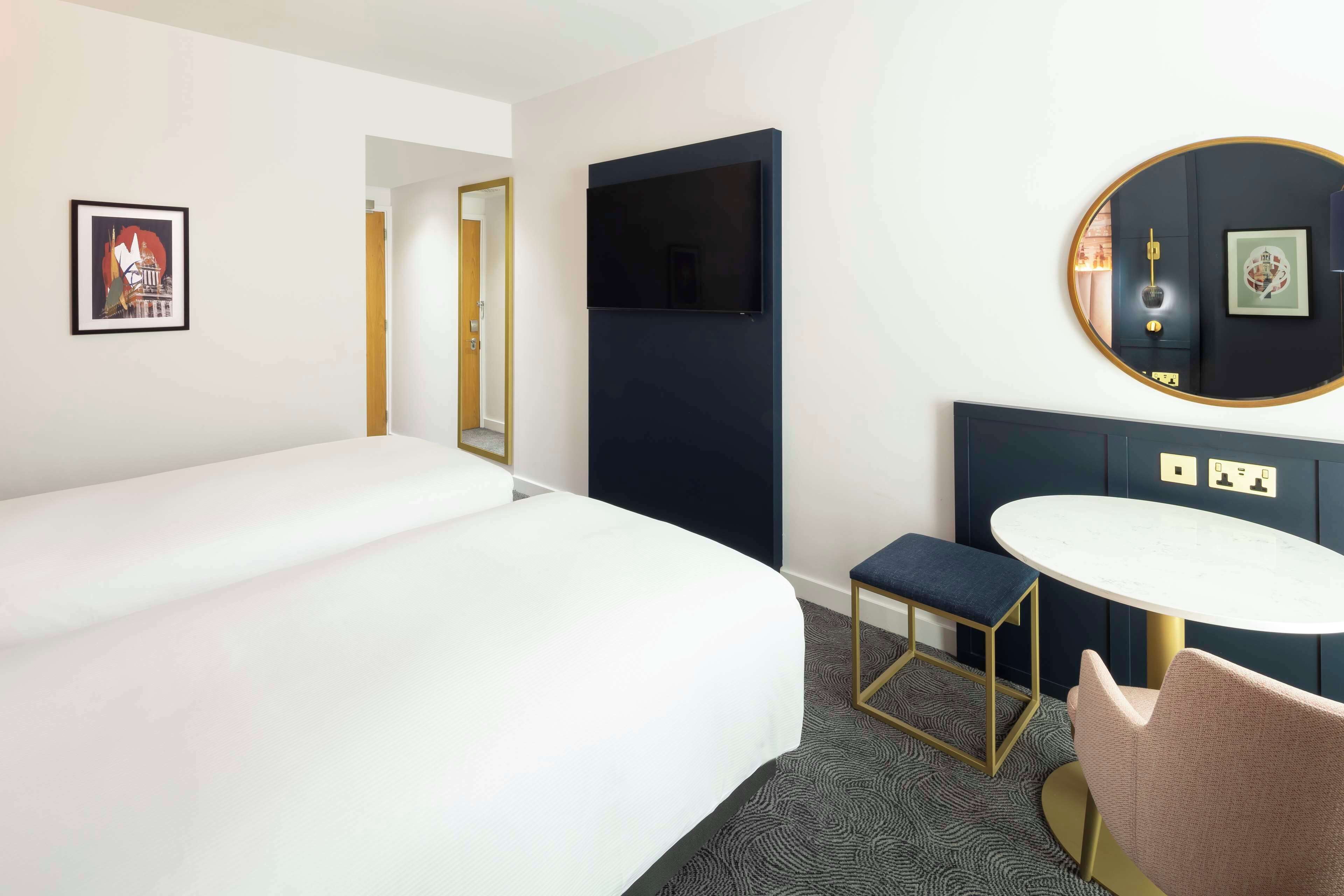 Habitación de hotel con una cama grande, una mesita redonda pequeña, un banco y un televisor instalado en la pared.
