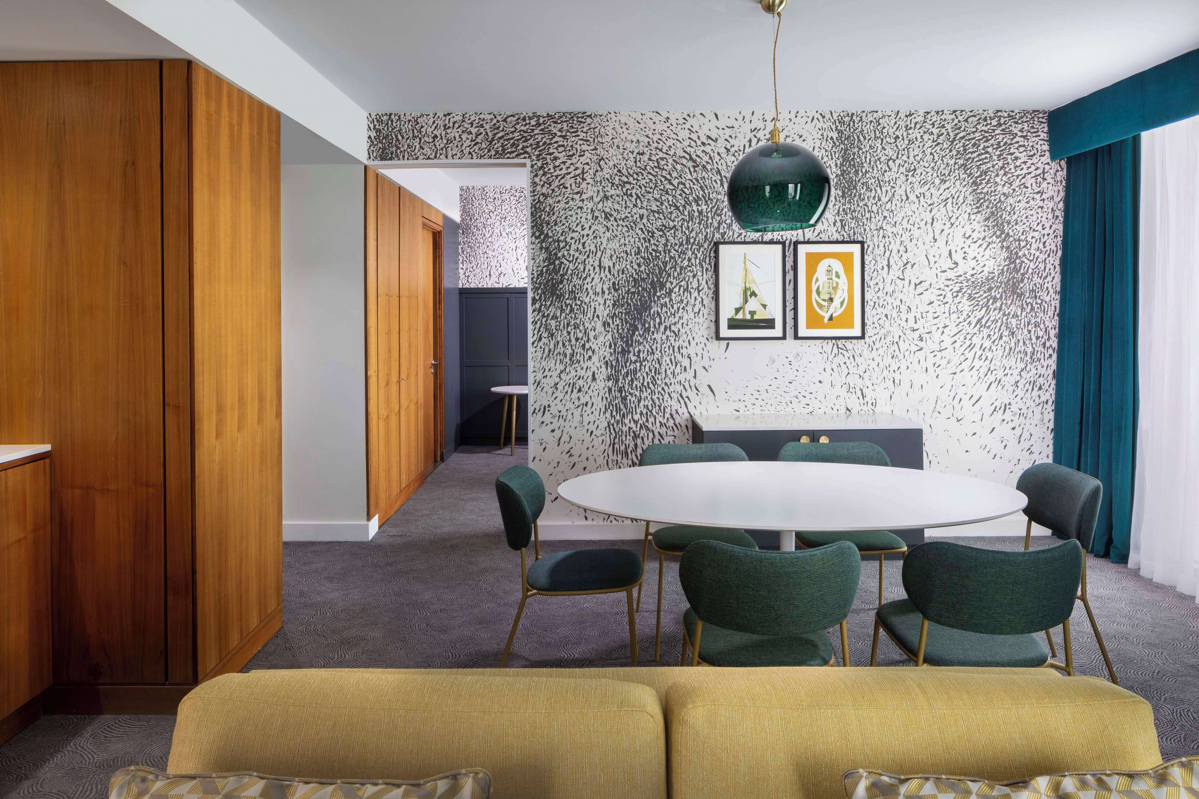 A modern dining area with a round table, green upholstered chairs, and a patterned wall.