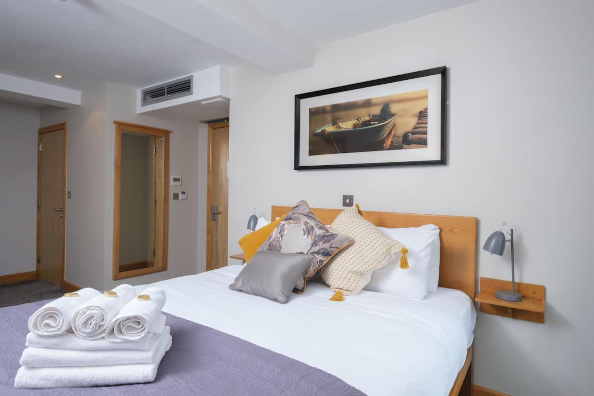 A neatly made bed with rolled towels, a framed picture of a boat, and a bedside table with a lamp.