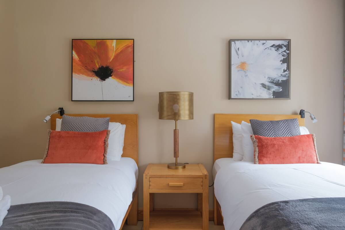A hotel room with two beds, each with white bedding and orange pillows. There are two nightstands with lamps between the beds.