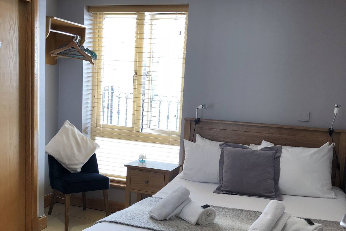 A neatly made bed with white linens and rolled towels, a wooden headboard, a bedside table with a lamp, and a window with blinds.