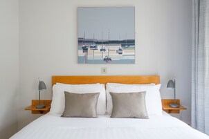 Una cama bien hecha con sábanas blancas y dos almohadas, un cabecero de madera y un cuadro con barcos en la pared.