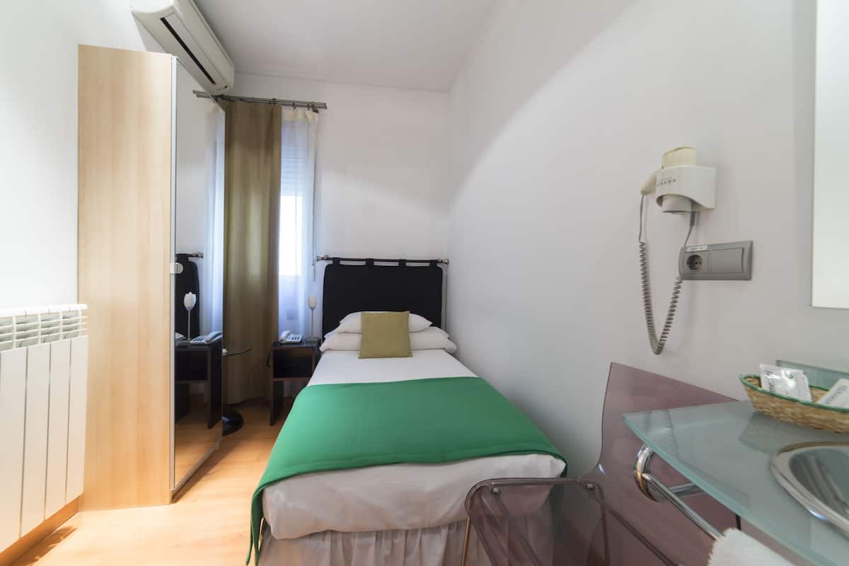 Quarto de hotel com cama, escrivaninha, cadeira, mesa de vidro, telefone e radiador.