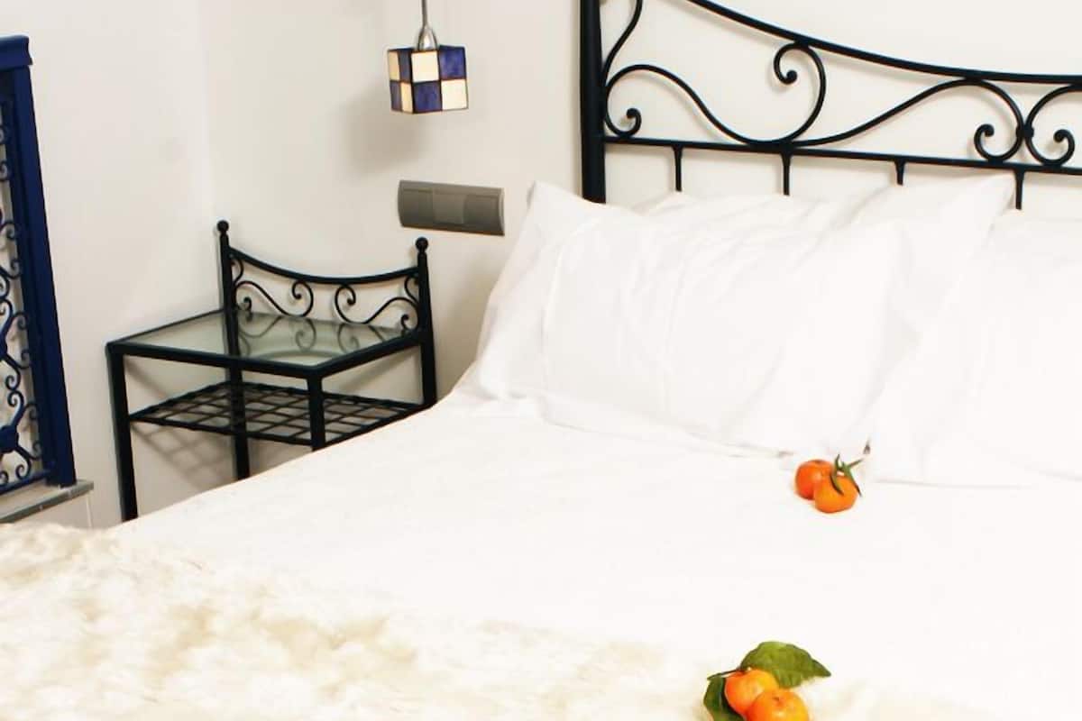 Uma cama com um cobertor branco e fofo, uma mesa pequena com laranjas e uma cadeira de metal.