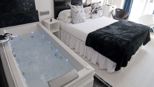 A modern bedroom with a large bathtub, a bed with white bedding and black throw, and a bedside table with a lamp. - Hostal Santo Domingo (Madrid)