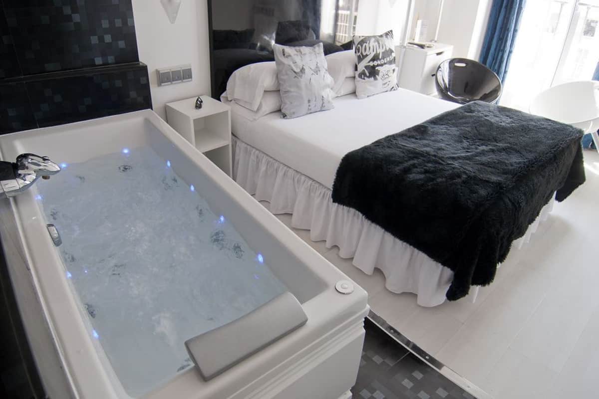 Quarto moderno com uma banheira grande, uma cama com roupa de cama branca e manta preta, e uma mesa de cabeceira com abajur.