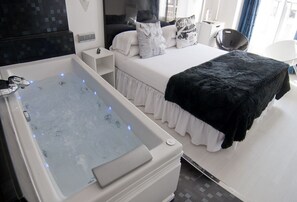 A modern bedroom with a large bathtub, a bed with white bedding and black throw, and a bedside table with a lamp. - Hostal Santo Domingo (Madrid)