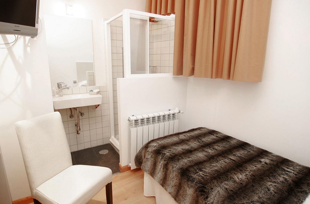 A small, compact room with a white chair, a bed with a fur blanket, a white radiator, and a shower area.