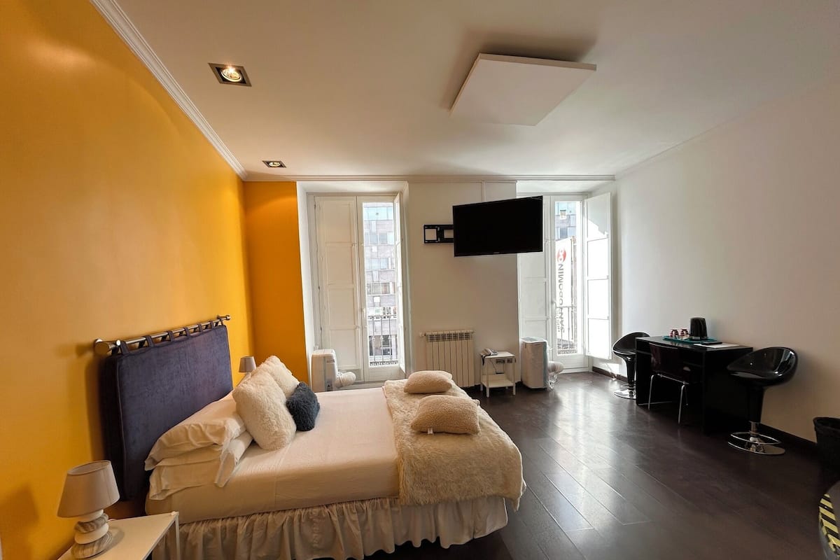 Quarto de hotel com cama, escrivaninha, cadeira, televisão e janela.