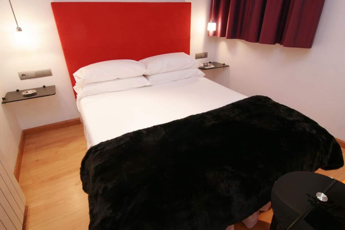 Quarto de hotel com cabeceira vermelha, roupa de cama branca, manta de pele preta, mesa de cabeceira e abajur.