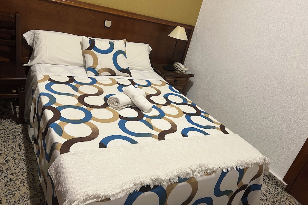 Una cama bien hecha con una colcha estampada, un plaid blanco y una toalla blanca enrollada.