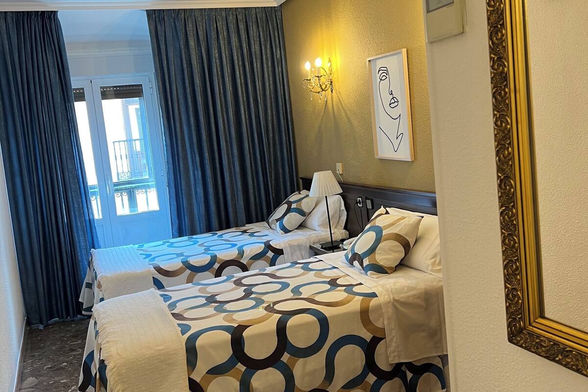 Habitación de hotel con dos camas, una araña y una obra de arte enmarcada, con vistas al exterior.