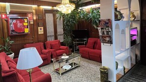 Un salón con sofás rojos, una mesa de centro de vidrio, una araña, un televisor y una estantería llena de revistas.