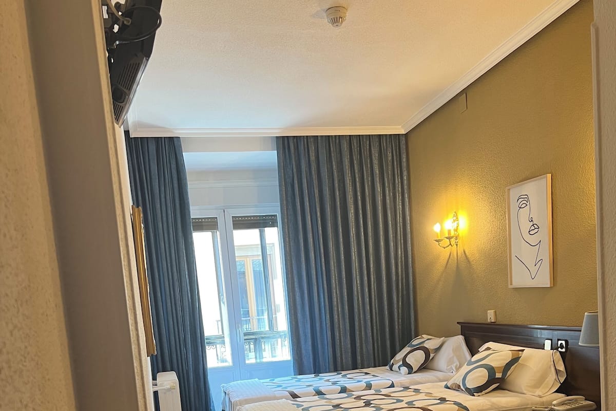 Habitación de hotel con dos camas, un escritorio con lámpara, una lámpara de pared y un ventanal con cortinas.