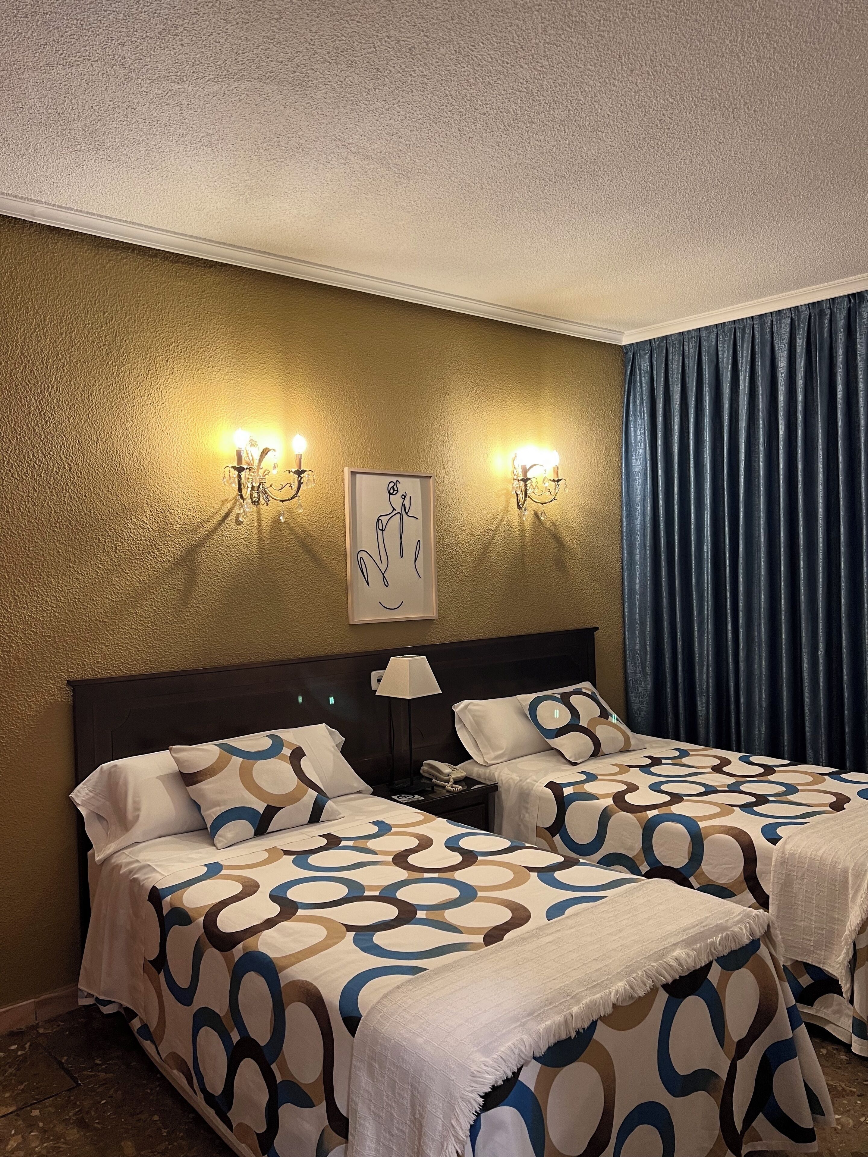A hotel room with two beds, a headboard, wall sconces, a framed artwork, and a curtain.