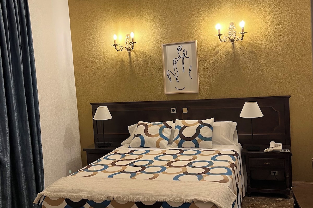 Una habitación de hotel con cama, dos lámparas de noche, mesita de noche con teléfono, y aplique de pared.