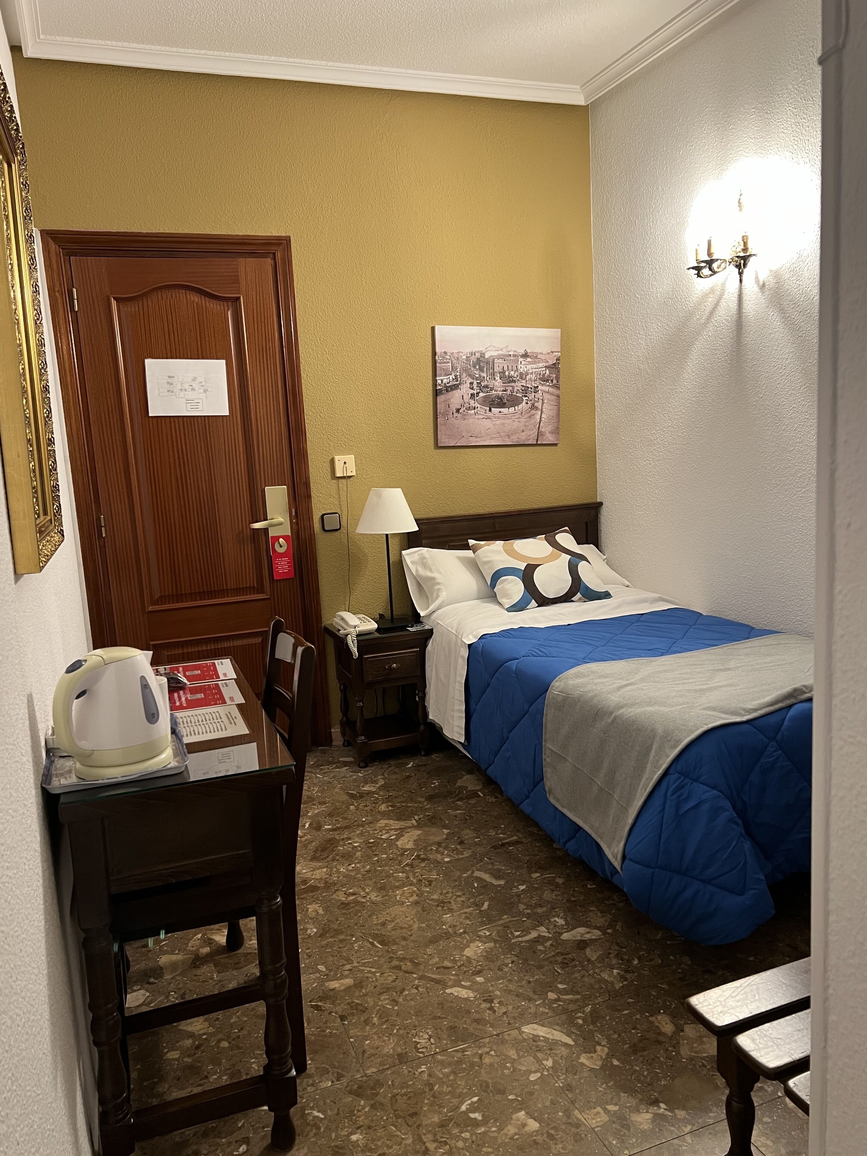 Quarto de hotel com porta de madeira, cama com roupa de cama azul, mesa de cabeceira com abajur e uma pequena mesa com chaleira e telefone.