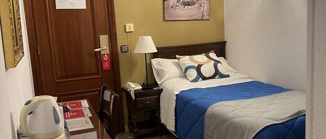Quarto de hotel com porta de madeira, cama com roupa de cama azul, mesa de cabeceira com abajur e uma pequena mesa com chaleira e telefone.
