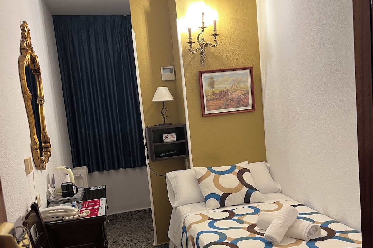 Habitación de hotel con cama, escritorio con teléfono y cafetera, una silla, una mesita y una lámpara de pared, además de un cuadro.