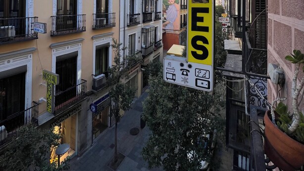 Un cartel de hostal con el nombre "HOSTAL SONSOLLES" en una calle estrecha flanqueada por edificios de varios pisos, balcones y un cielo azul despejado.