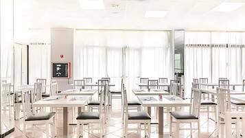 Un comedor amplio con mesas y sillas blancas, un detector de incendios y ventanas grandes con cortinas.