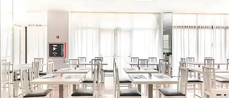 A spacious dining area with white tables and chairs, a fire alarm, and large windows with curtains.