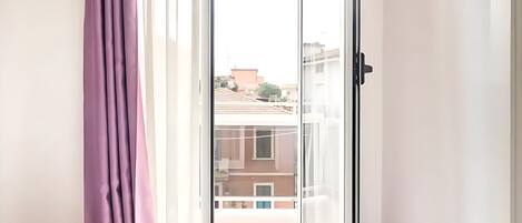 A room with a bed, a window with purple curtains, and a view of buildings outside.