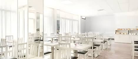 A spacious, well-lit dining area with white tables and chairs, large windows, and a clean, modern design.