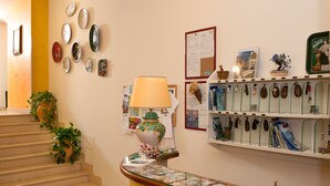 A reception area with a counter, a lamp, a plant, a bulletin board, and a display of keychains. - Hotel De Rosa (Maiori)